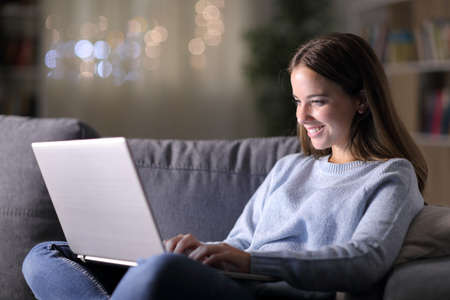 Happy woman using laptop sitting on a couch in the living room in the night at homeの写真素材