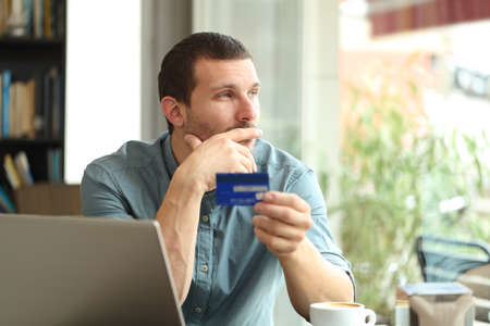 Pensive man holding a credit card thinking looking away through a window sitting in a coffee shopの写真素材
