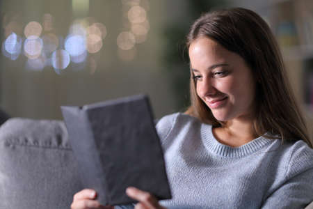 Relaxed woman reading an ebook sitting on a couch in the night at homeの写真素材