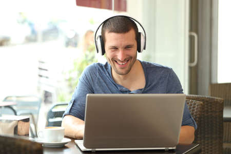 Happy man with headphones watching media on laptop sitting in a coffee shopの写真素材