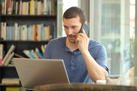 Serious man talks on phone and uses a laptop sitting at home or coffee shopの写真素材