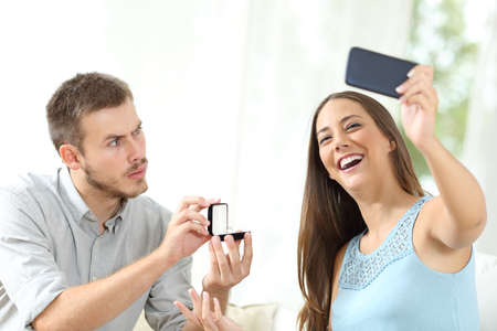 Upset man proposing marriage and girlfriend taking selfie sitting on a couch at homeの写真素材