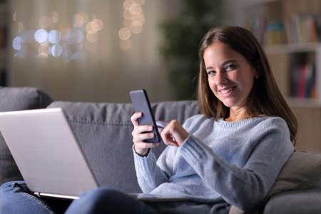 Happy woman in the night with multiple devices looks at you sitting on a couch in the night at homeの写真素材