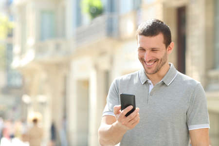 Happy adult man checking mobile phone content walking in the streetの写真素材