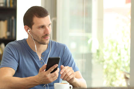 Relaxed adult man listening to music from smart phone looking through a window sitting in a coffee shop or homeの写真素材