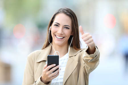 Front view portrait of a happy girl listening to music wearing earbuds and holding smart phone posing with thumbs up in the streetの写真素材