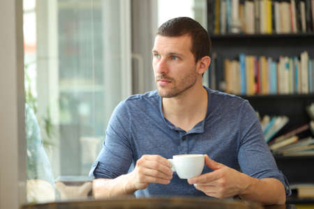 Pensive man holding coffee looking at side through a window sitting in a barの写真素材