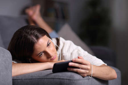 Relaxed girl watching and listening movie on phone lying on a couch in the night at homeの写真素材