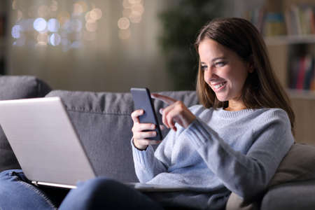 Happy woman using laptop checks smart phone sitting on a couch in the night at homeの写真素材