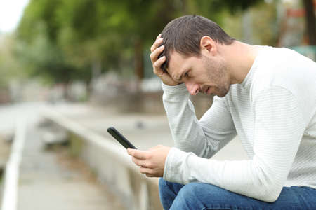Side view portrait of sad adult man checking bad news on smart phone in a parkの写真素材
