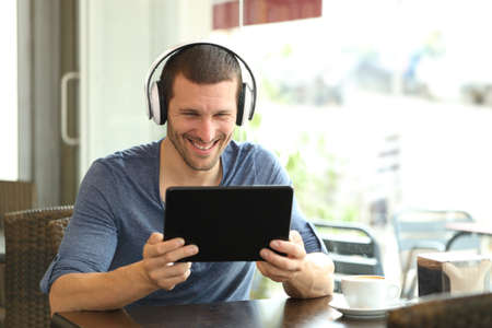 Happy man with headphones using a tablet sitting in a coffee shopの写真素材