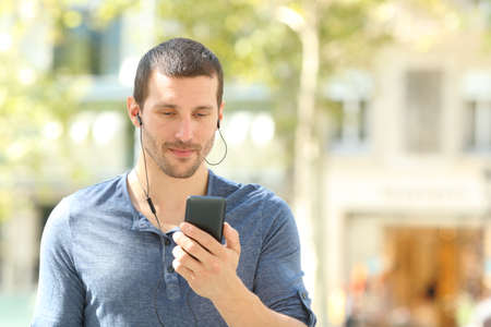 Front view of an adult man listening to music checking cell phone walking in the streetの写真素材