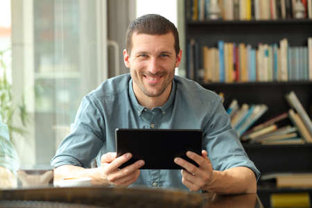 Front view portrait of a happy man holding tablet looking at camera sitting in a coffee shop or homeの写真素材