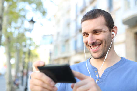 Happy adult man wearing earbuds watching videos on horizontal mobile phone in the streetの写真素材