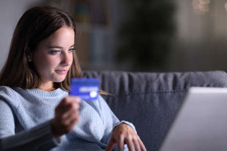 Serious woman using laptop and credit card to buy online sitting on a couch in the living room in the night at homeの写真素材
