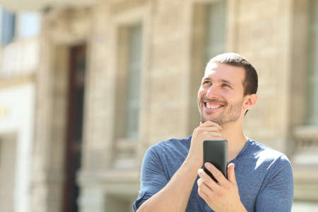 Pensive man holding mobile phone looks at side wondering standing in the streetの写真素材