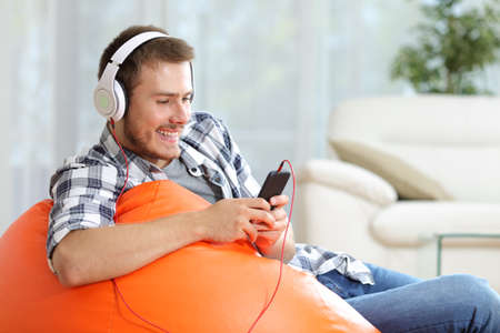 Happy man listening to music sitting on an orange pouf at homeの写真素材
