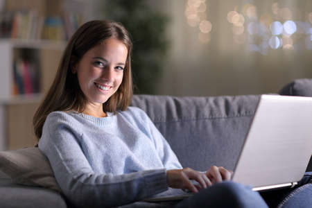 Happy woman using laptop looking at camera sitting on a couch in the living room in the night at homeの写真素材
