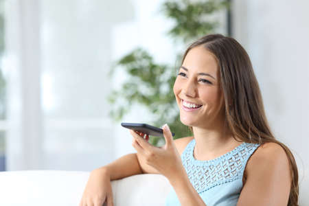 Happy woman uses voice recognition on mobile phone sitting on a couch at homeの写真素材