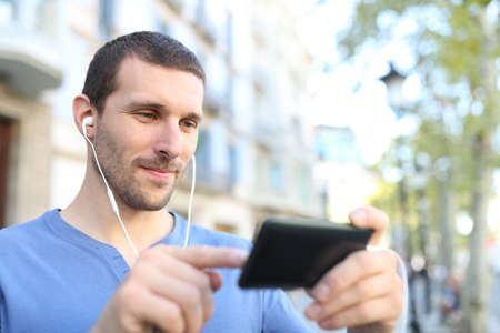 Adult man wearing earphones searching video on horizontal mobile phone in the streetの写真素材