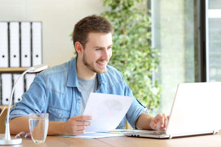 Casual happy entrepreneur working using laptop holding document comparing data at office