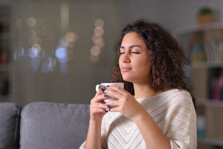 Relaxed woman smelling coffee cup sitting on a couch in the night at homeの写真素材