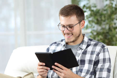Man wearing eyeglasses reading an ebook sitting on a couch in the living room at homeの写真素材