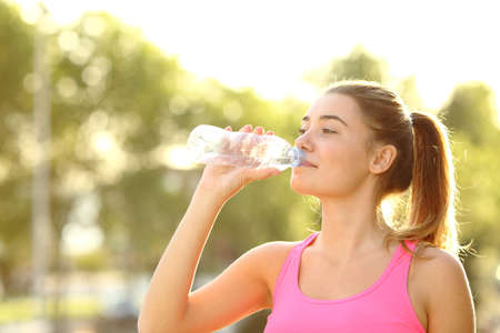 Happy runner drinking water from a bottle standing in a park after sport