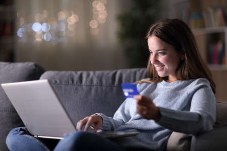 Happy woman in the night buying on line with credit card and laptop sitting on a couch at homeの写真素材