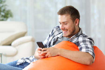 Happy man sitting on an orange pouf at home using mobile phoneの写真素材