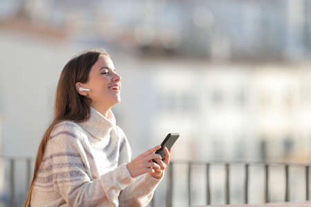 Happy woman listens to music with wireless earbuds and smart phone in a balcony in winter