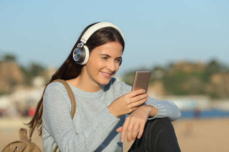 Happy girl listens to music with headphones and mobile phone sitting in a coast town beachの写真素材