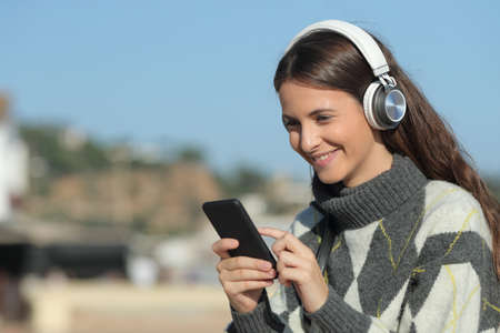 Happy girl listening to music using mobile phone and headphones in winter sitting in a coast town beachの写真素材