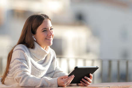 Happy woman listening to music with a tablet and wireless earphones looks away in a balcony in winterの写真素材