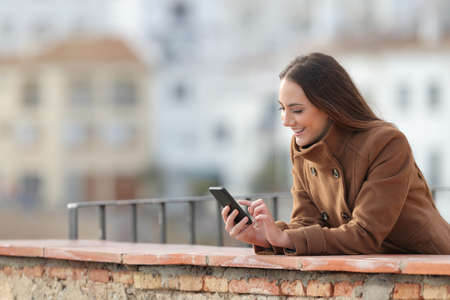 Happy woman using a mobile phone in winter in a balcony in a rural townの写真素材