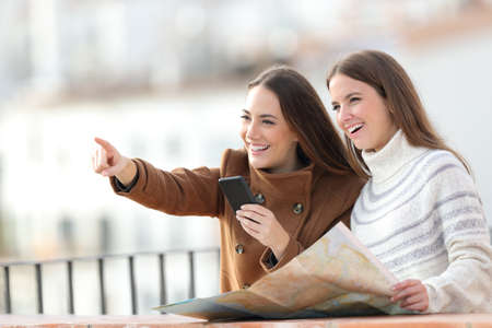 Two happy tourists holding paper map and mobile phone pointing away in a balcony on winter holidayの写真素材