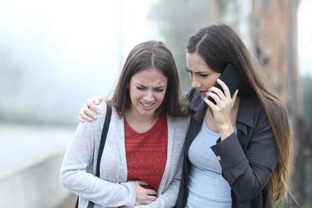 Front view portrait of a girl suffering belly ache and a friend calling assistanceの写真素材