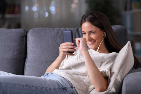 Happy teenage girl wearing earbuds listening to music on mobile phone sitting on a couch in the night at homeの写真素材