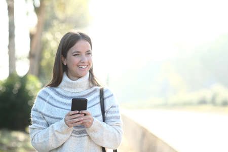 Front view portrait of a happy girl wearing sweater holding smart phone looking away in a parkの写真素材