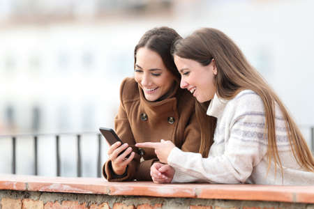 Two happy friends checking mobile phone in winter in a balconyの写真素材