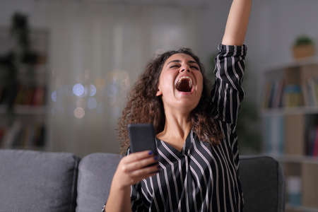 Excited elegant woman holding smart phone sitting on a couch in the night at homeの写真素材