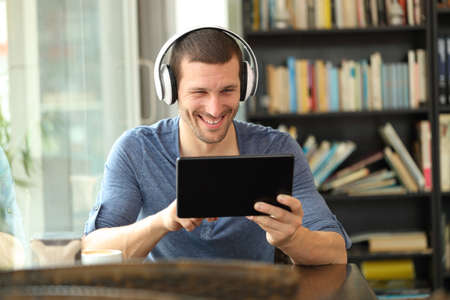 Front view of happy man with headphones using a tablet sitting in a coffee shop or homeの写真素材