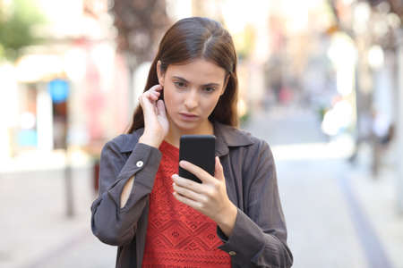 Front view of a serious girl using mobile phone touching hair walking in the streetの写真素材