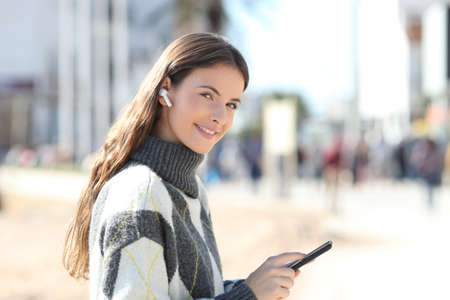 Happy girl holds mobile phone listening to music looking at camera standing in the street in winterの写真素材