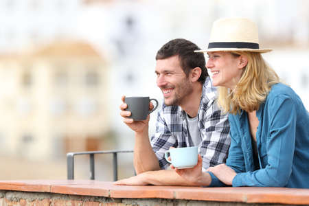 Happy couple of tourists drinking coffee and looking away in a balcony in a rural town on vacationの写真素材