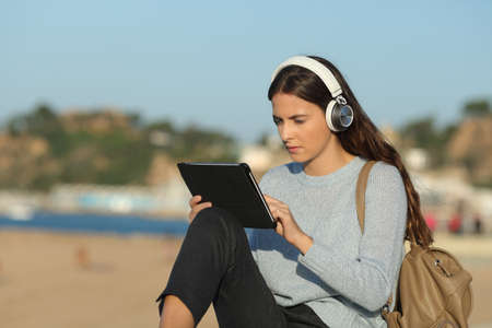 Serious girl e-learning with a tablet and headphones sitting in a coast town beachの写真素材