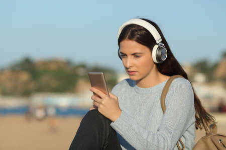 Serious girl listening to music checking smart phone sitting in a coast town beachの写真素材