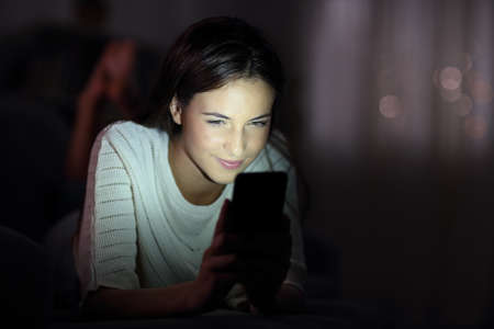 Serious girl using smart phone in the night with the face lit lying on a couch in the night at homeの写真素材
