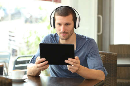 Serious man with headphones using a tablet sitting in a coffee shopの写真素材