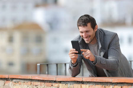Happy adult man in winter using mobile phone standing in a balcony outdoors in a rural townの写真素材